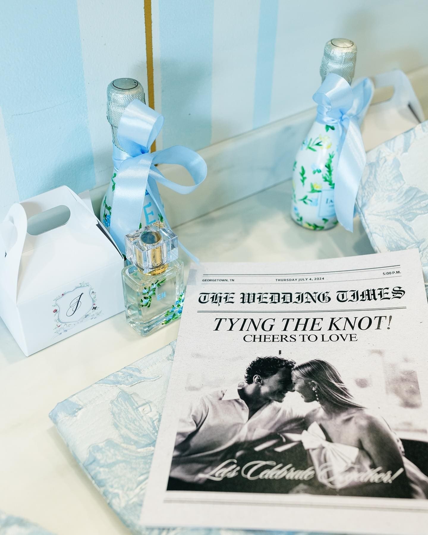 Flat lay of elegant wedding welcome gifts featuring custom champagne bottles, personalized perfume, favor boxes, and a wedding newspaper on a soft blue patterned fabric backdrop.